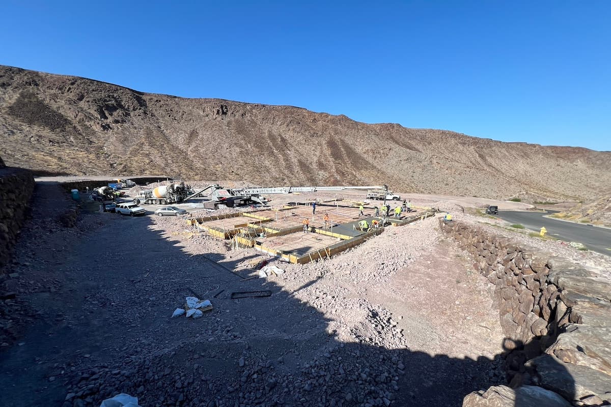 Construction site on a desert hillside with workers pouring a concrete foundation near a mountain road.