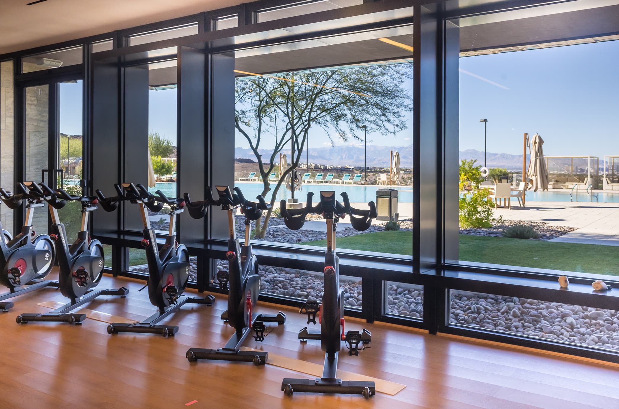 A row of stationary bikes inside the clubhouse fitness center, with floor-to-ceiling windows overlooking the resort-style pool, desert landscaping, and mountain views.