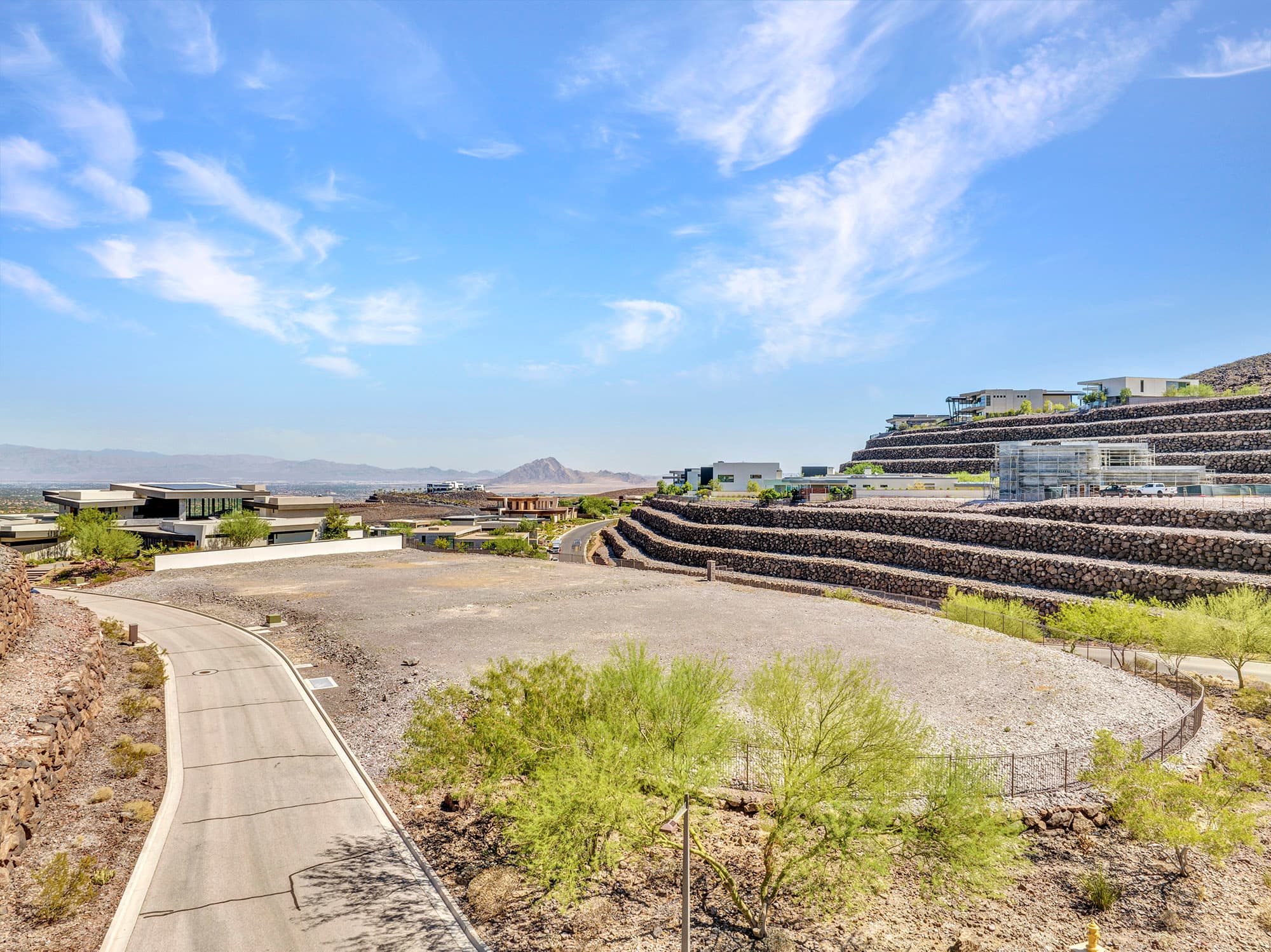 Elevated view of a large homesite at Ascaya surrounded by signature terraced rockery walls, modern custom residences, desert landscaping, and distant mountain views under a dramatic blue sky.