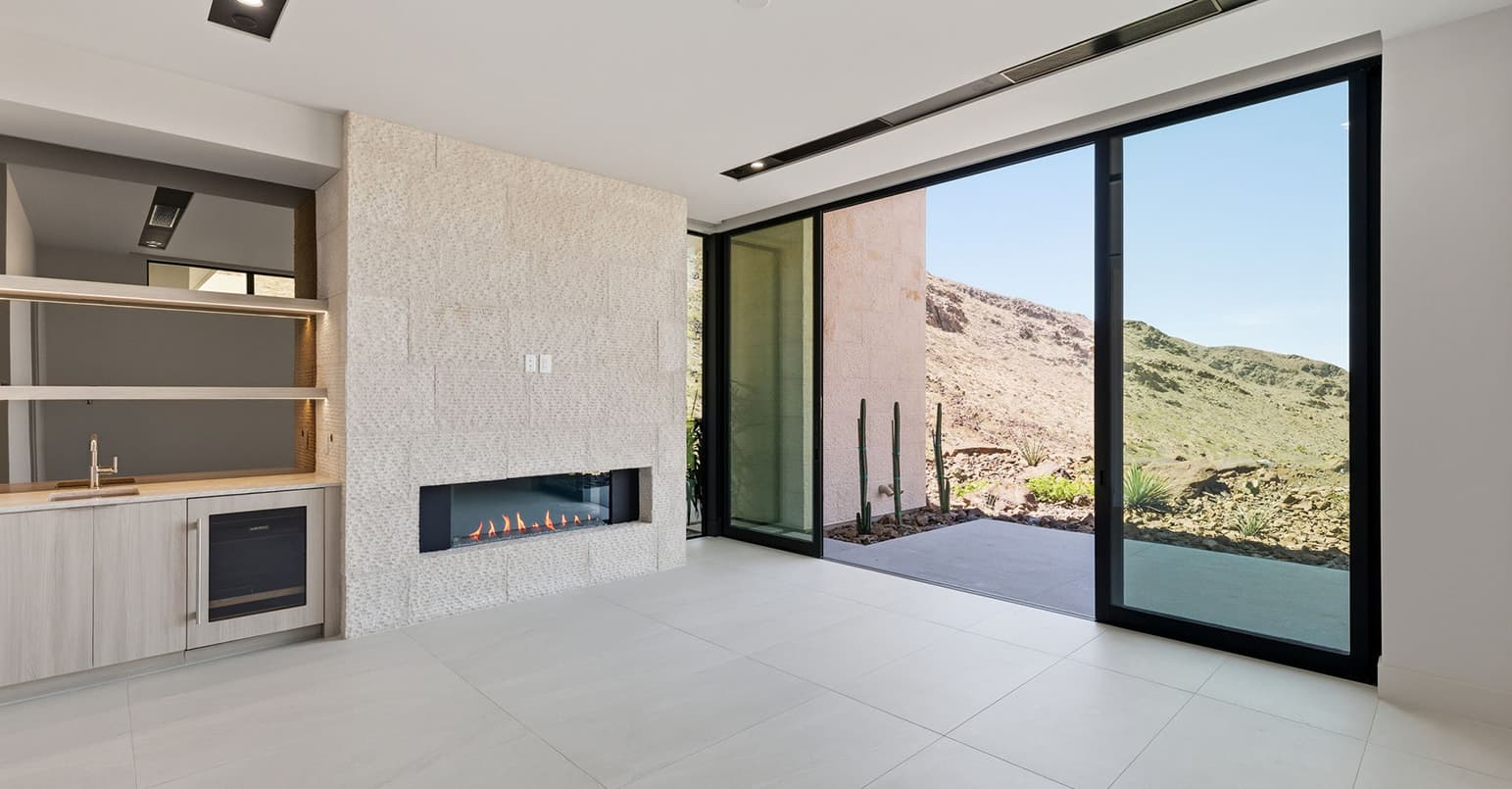 Modern indoor-outdoor living space with linear fireplace, built-in wet bar, floor-to-ceiling sliding glass doors, and expansive desert mountain views
