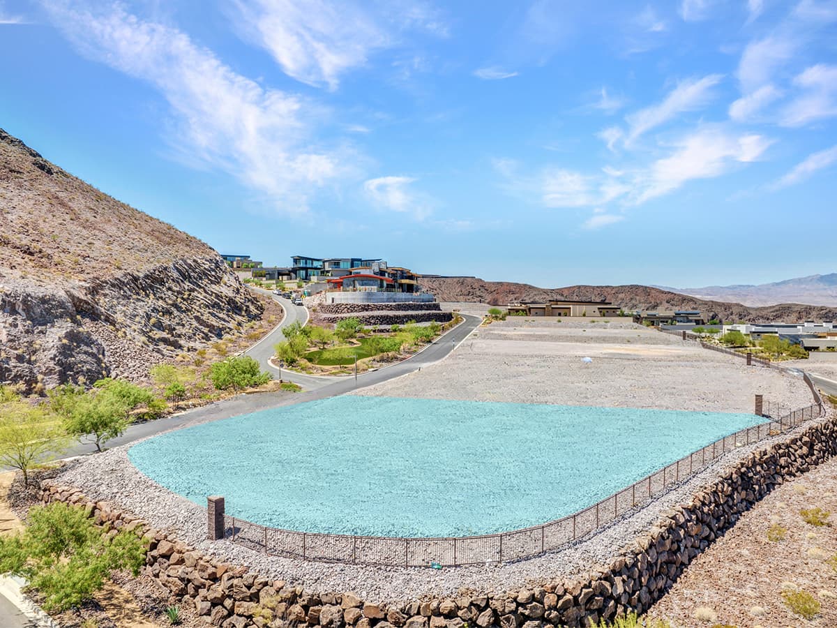 Elevated desert residential lot with retaining wall, scenic mountain views, and modern homes along a winding road under blue sky.