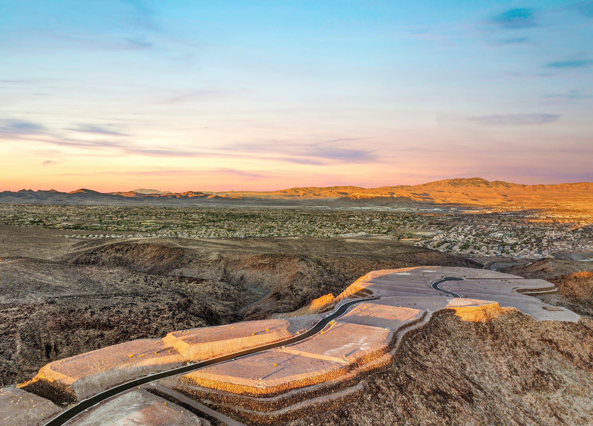 Aerial view of a desert hillside lot with a circular driveway pad overlooking a sprawling suburban city at sunset, with mountains in the distance.