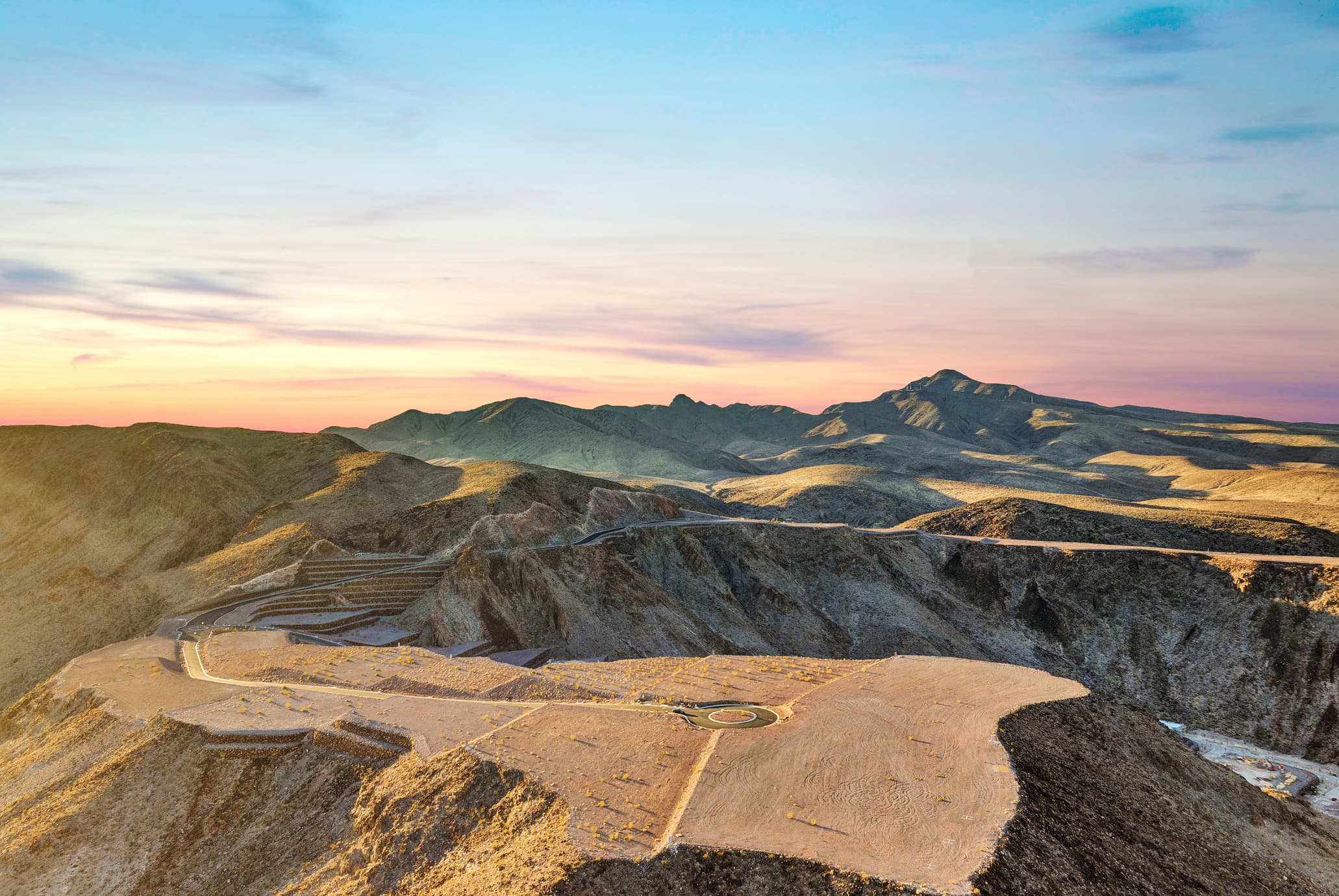 Aerial view of a desert hillside lot with a circular driveway pad overlooking a sprawling suburban city at sunset, with mountains in the distance.