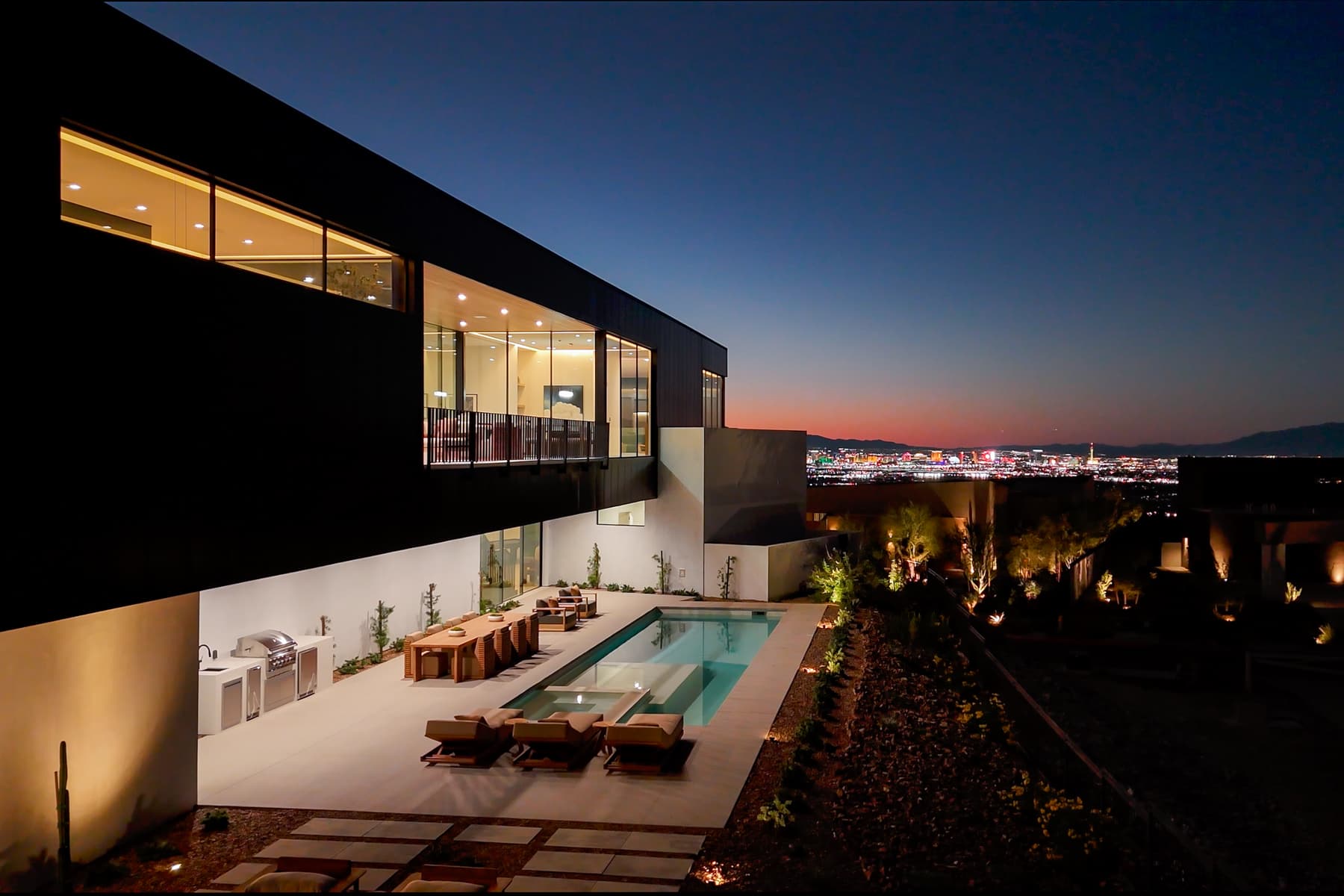 Modern luxury home at dusk with outdoor pool, patio seating, and city skyline views illuminated under a twilight sky.
