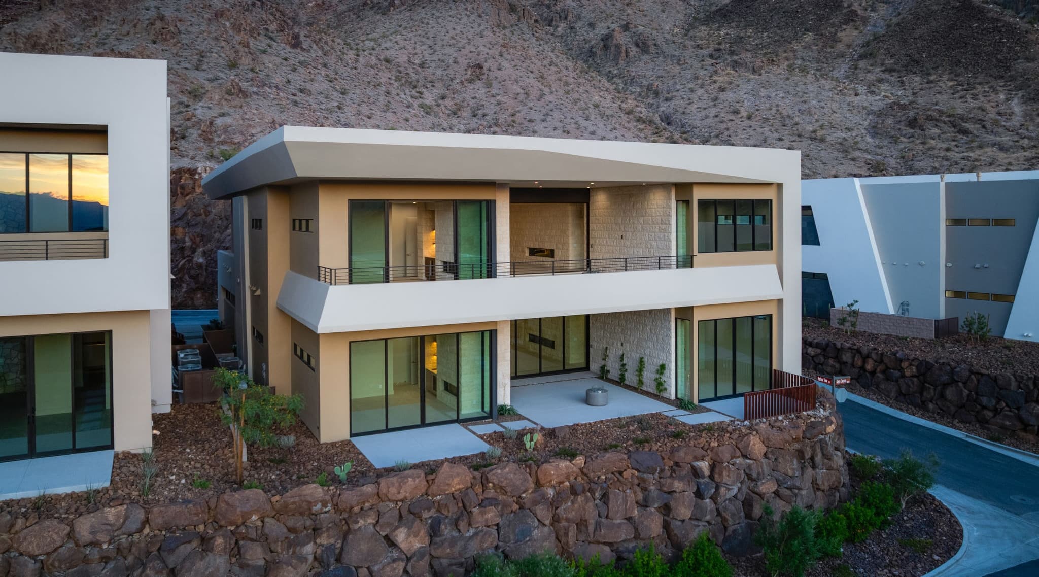 Modern two-story luxury residence with expansive glass walls, covered balconies, and contemporary desert architecture set into a rocky mountainside community at dusk.