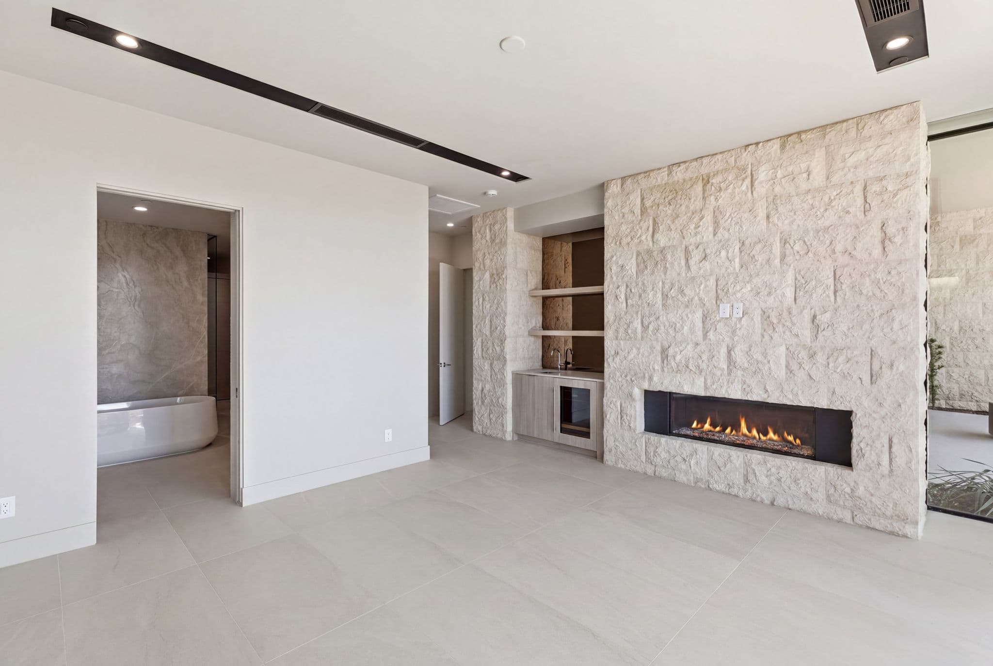 Primary bedroom retreat with linear fireplace, custom wet bar, textured stone accent wall, and direct access to a spa-inspired bathroom