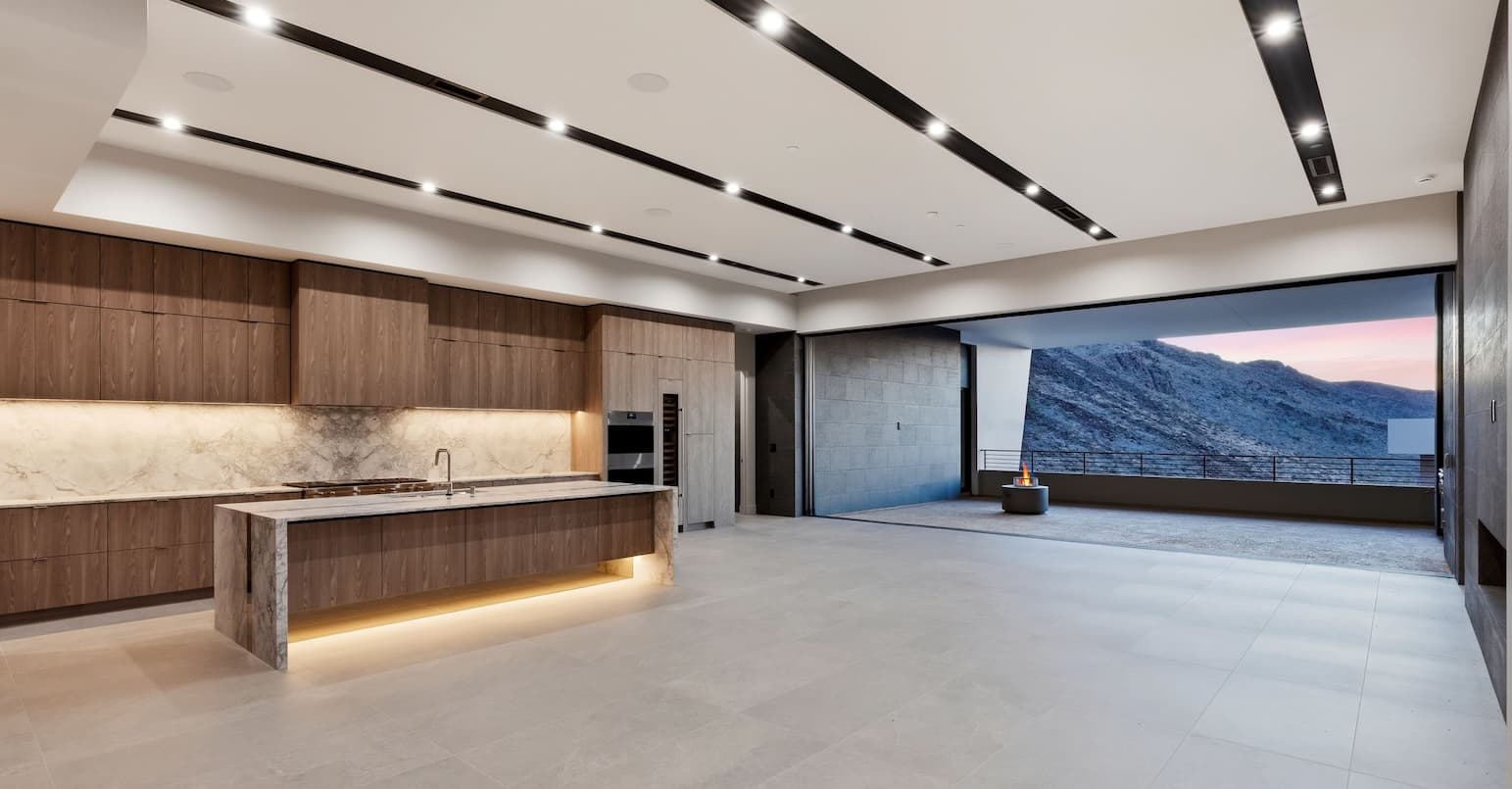 Modern luxury great room and kitchen with walnut cabinetry, marble waterfall island, recessed linear lighting, and expansive sliding glass doors opening to a covered terrace with panoramic mountain views at dusk.