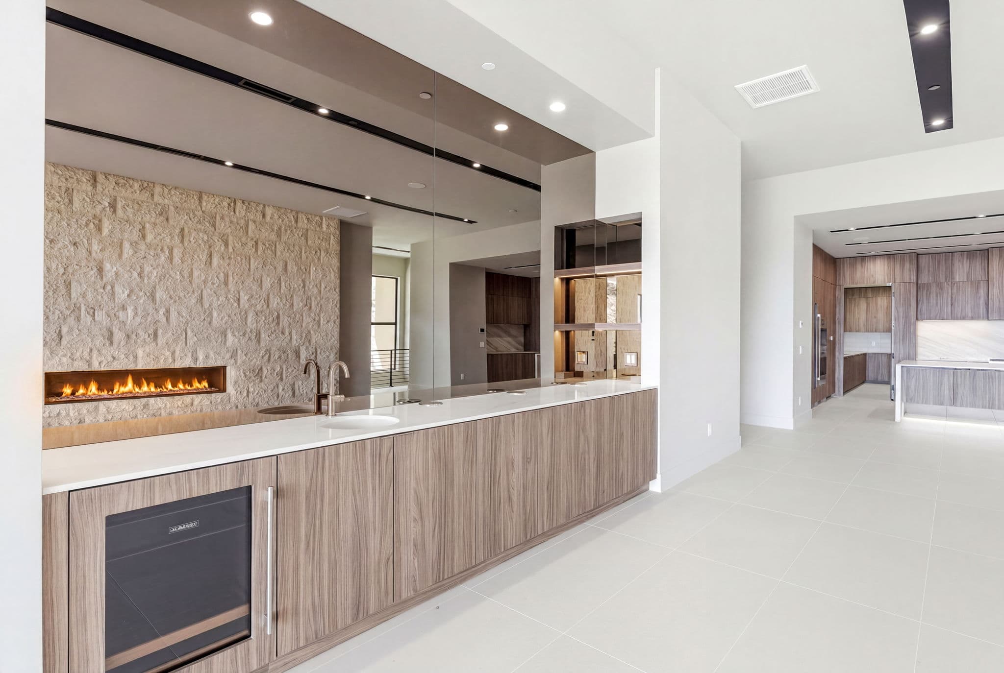 Modern entertainment bar with wood cabinetry, mirrored backsplash, under-counter beverage refrigerator, and linear stone fireplace in an open-concept interior