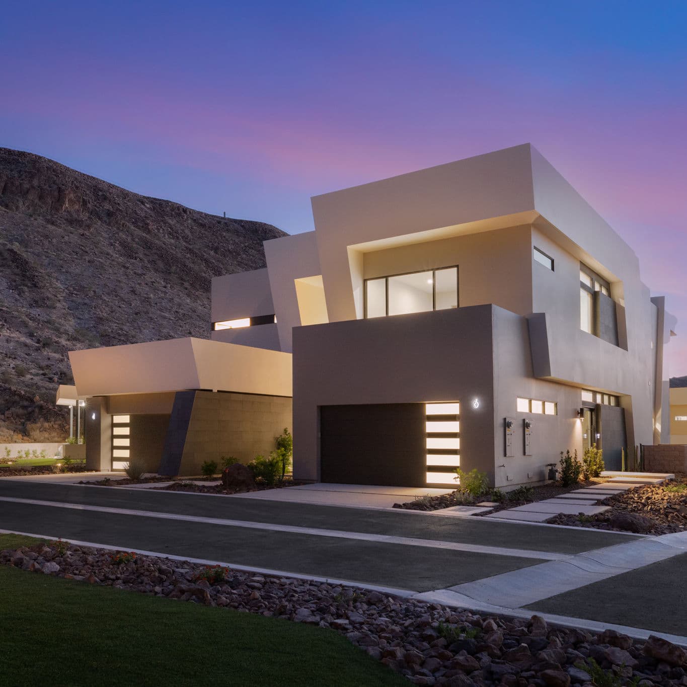 Modern desert-inspired residence with bold geometric architecture, illuminated garage doors, and minimalist landscaping set against a canyon backdrop at dusk