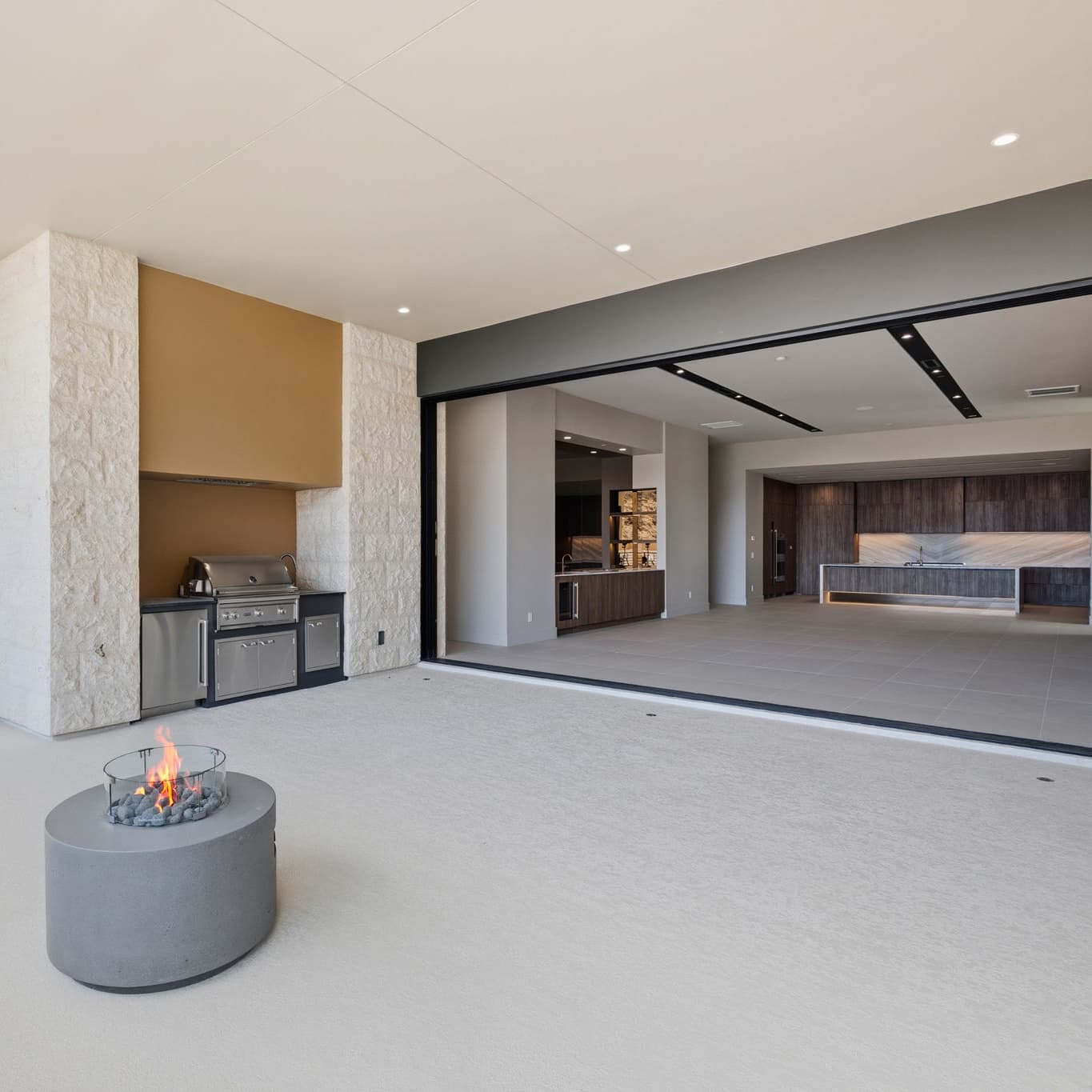 Expansive indoor-outdoor living space with retractable glass wall, modern outdoor kitchen, minimalist fire feature, and open-concept chef’s kitchen