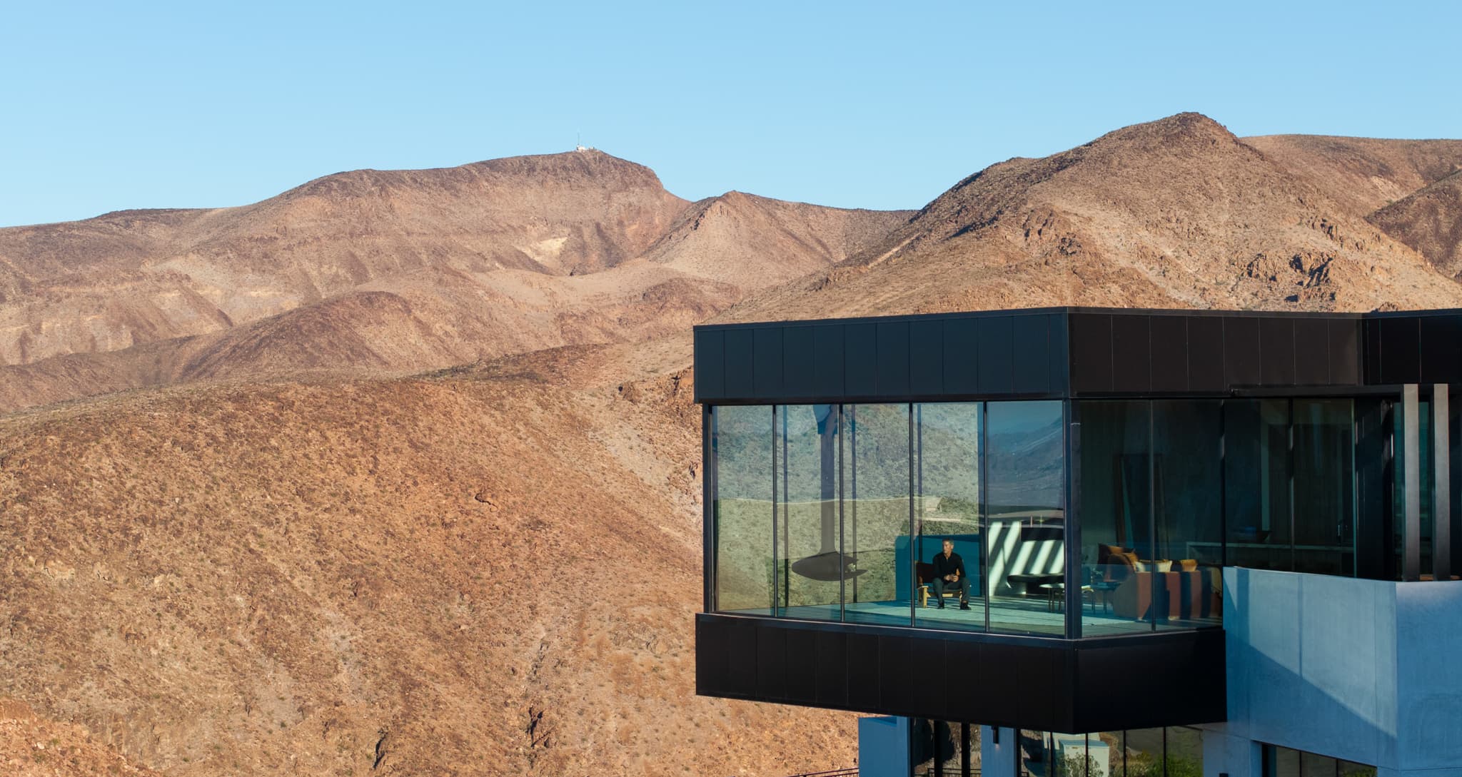 Modern cliffside luxury home with floor-to-ceiling glass walls overlooking rugged desert mountains, highlighting panoramic views and contemporary architecture.