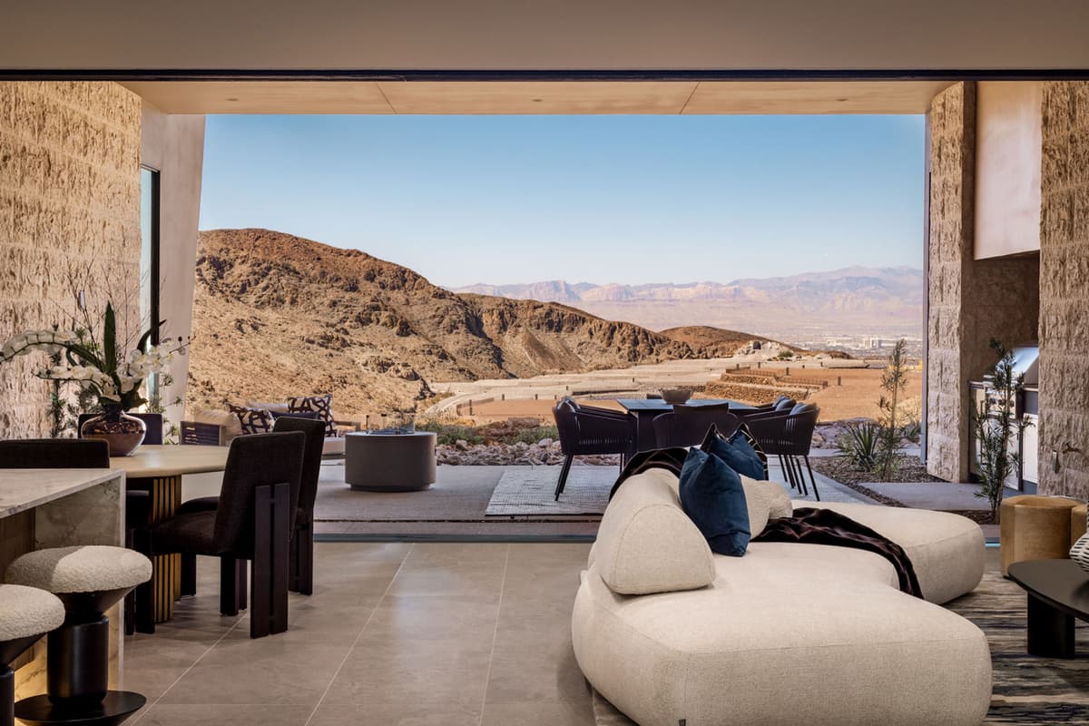 Elegant indoor-outdoor living space with a curved sofa and dining area, opening to a covered terrace with lounge seating, outdoor kitchen, and sweeping views of layered desert mountains and the valley beyond.
