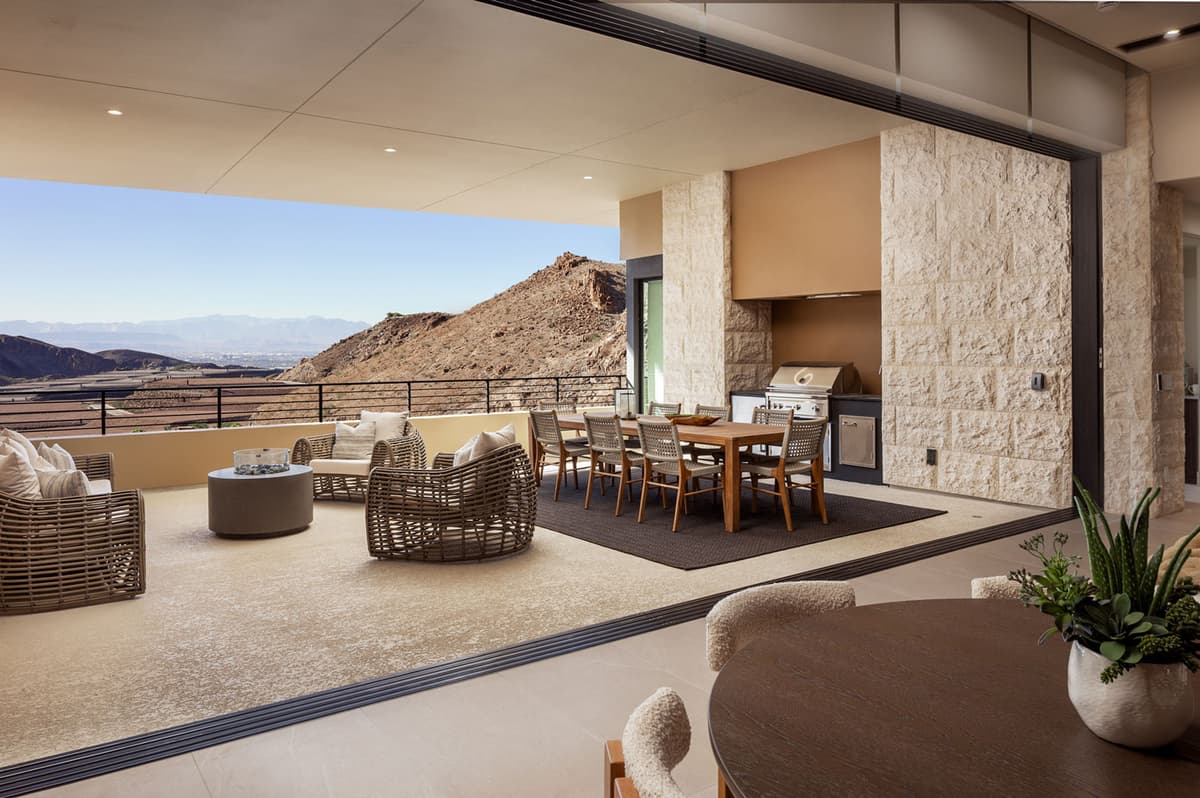 Seamless indoor-outdoor living space with a covered terrace featuring lounge seating, dining area, and built-in grill, all overlooking layered desert hills and expansive valley views.