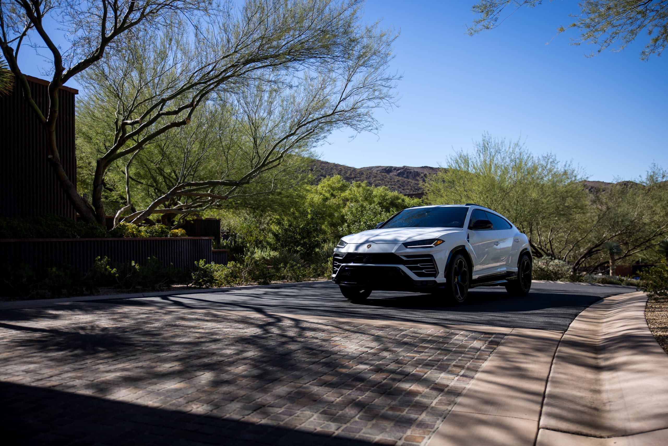 White luxury SUV driving through a modern desert community with landscaped greenery, mountain backdrop, and sunlit residential streets.