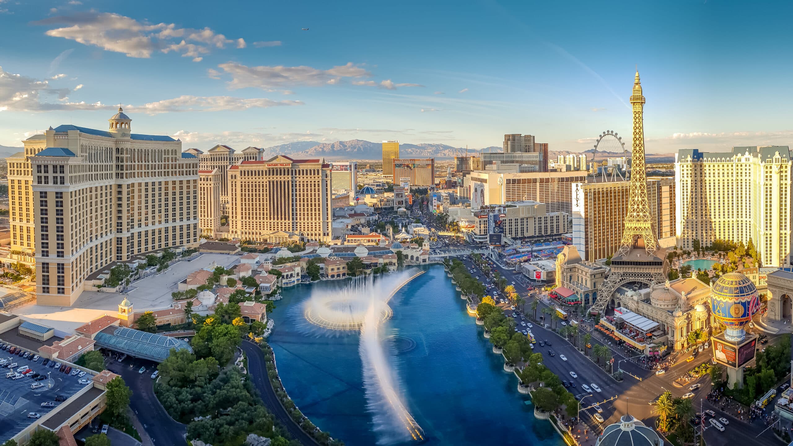 Aerial view of the Las Vegas Strip featuring Bellagio fountains in motion, Paris Las Vegas Eiffel Tower replica, and surrounding resort hotels under a clear daytime sky.