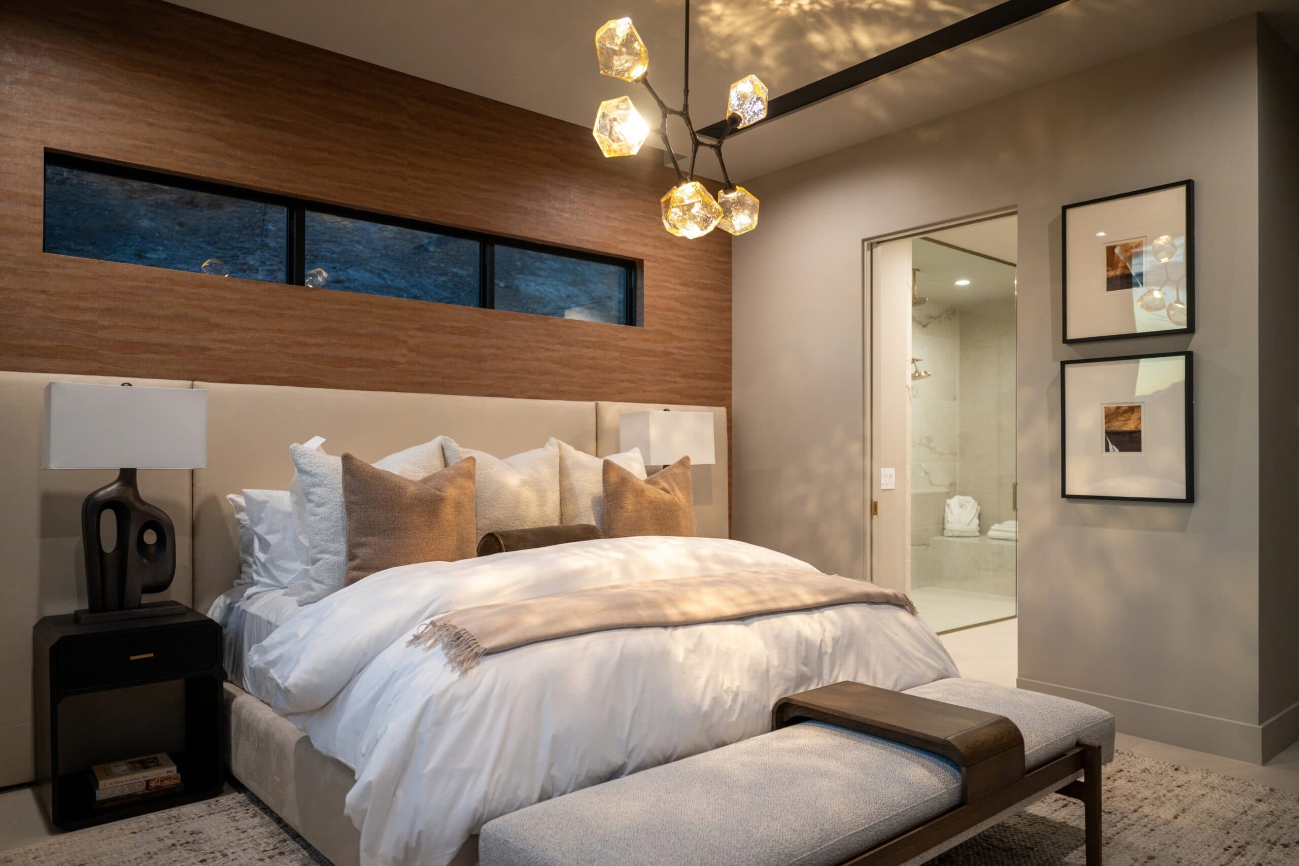 Luxury modern bedroom with warm wood accents, plush neutral bedding, statement lighting, and an ensuite bathroom.