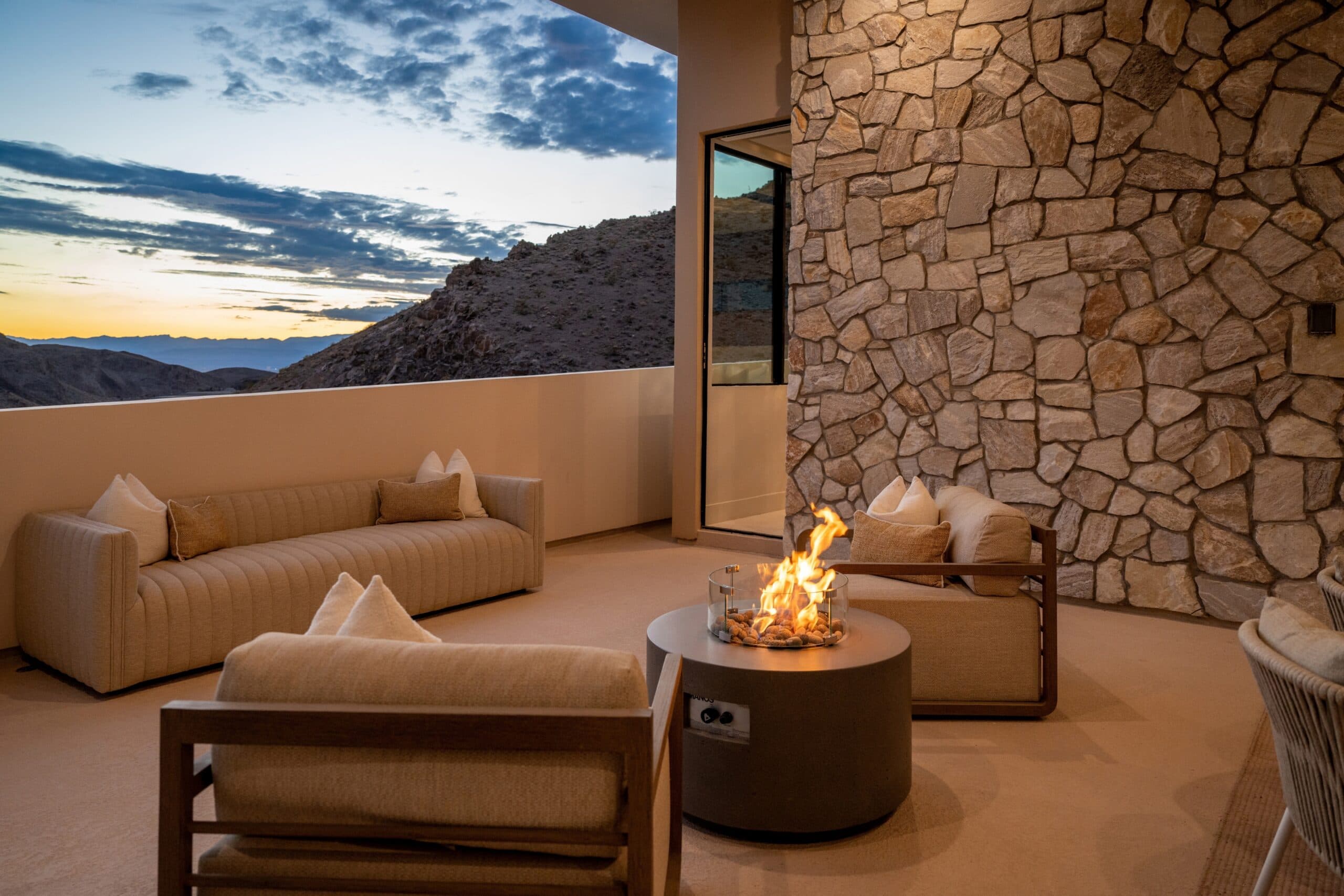 Luxury covered outdoor lounge with a modern fire pit, stone accent wall, and dramatic mountain sunset views.