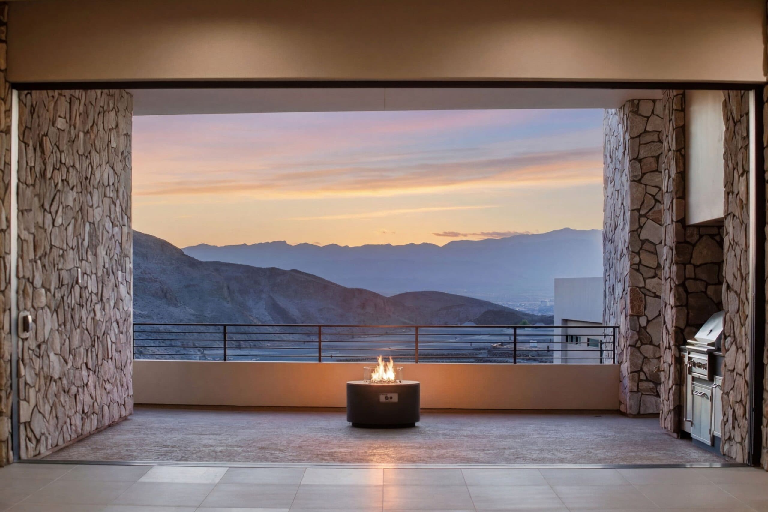 Luxury covered outdoor living space with stone walls, a fire pit, built-in grill, and panoramic mountain views at sunset.