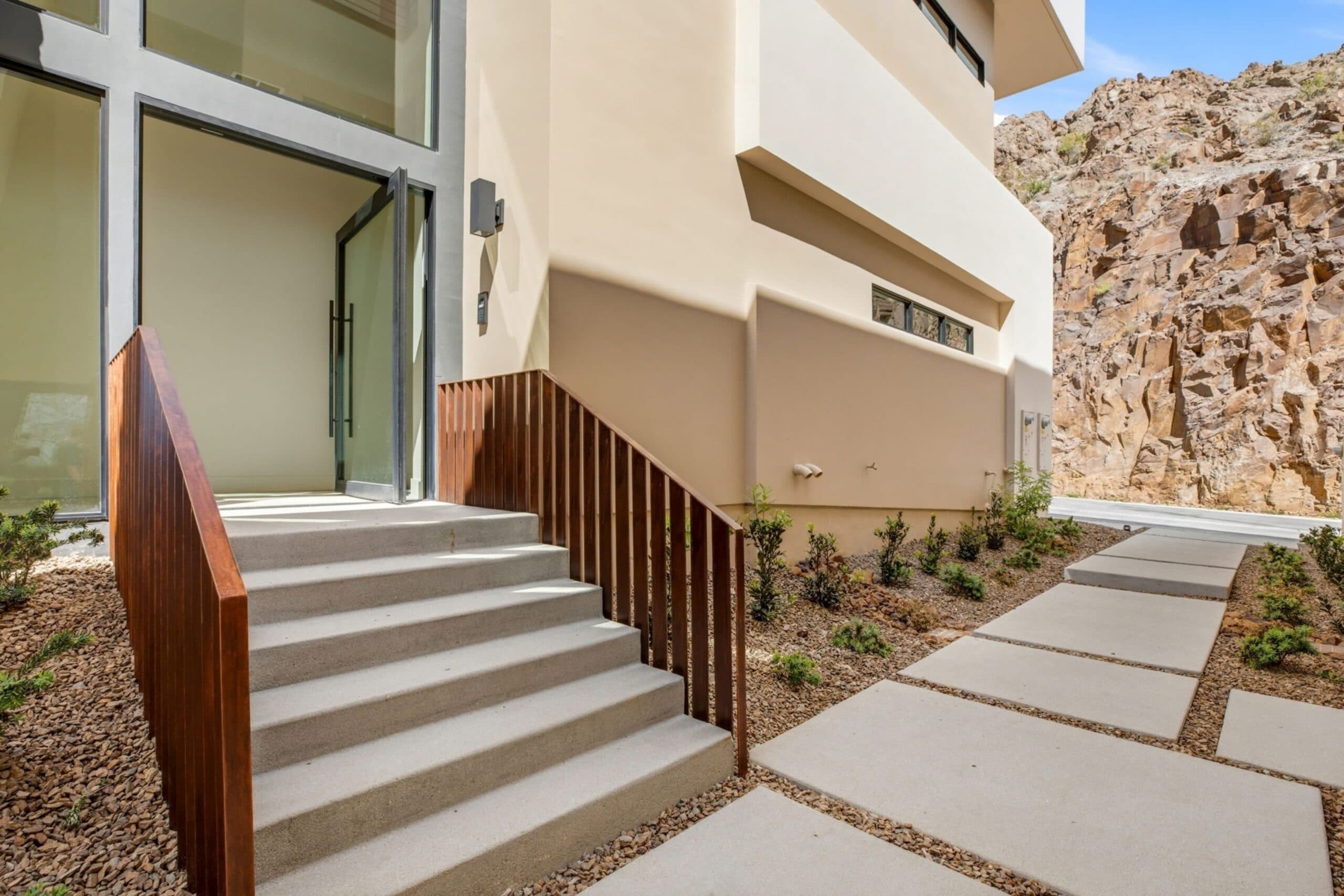 Modern luxury home entrance with concrete steps, warm wood railings, desert landscaping, and a dramatic rocky hillside backdrop.