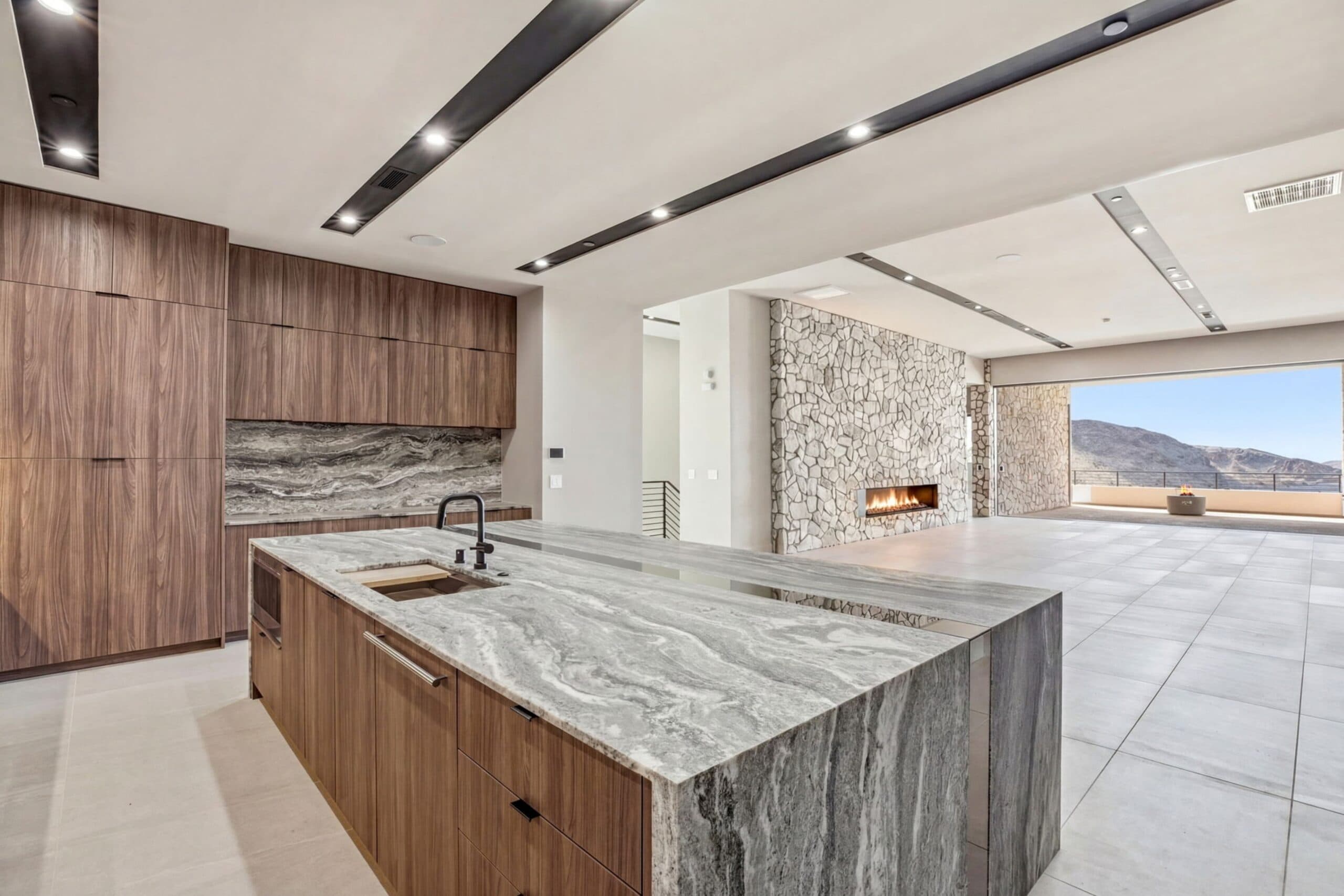 Modern luxury kitchen with rich wood cabinetry, a waterfall stone island, linear lighting, a stone fireplace, and expansive mountain views.