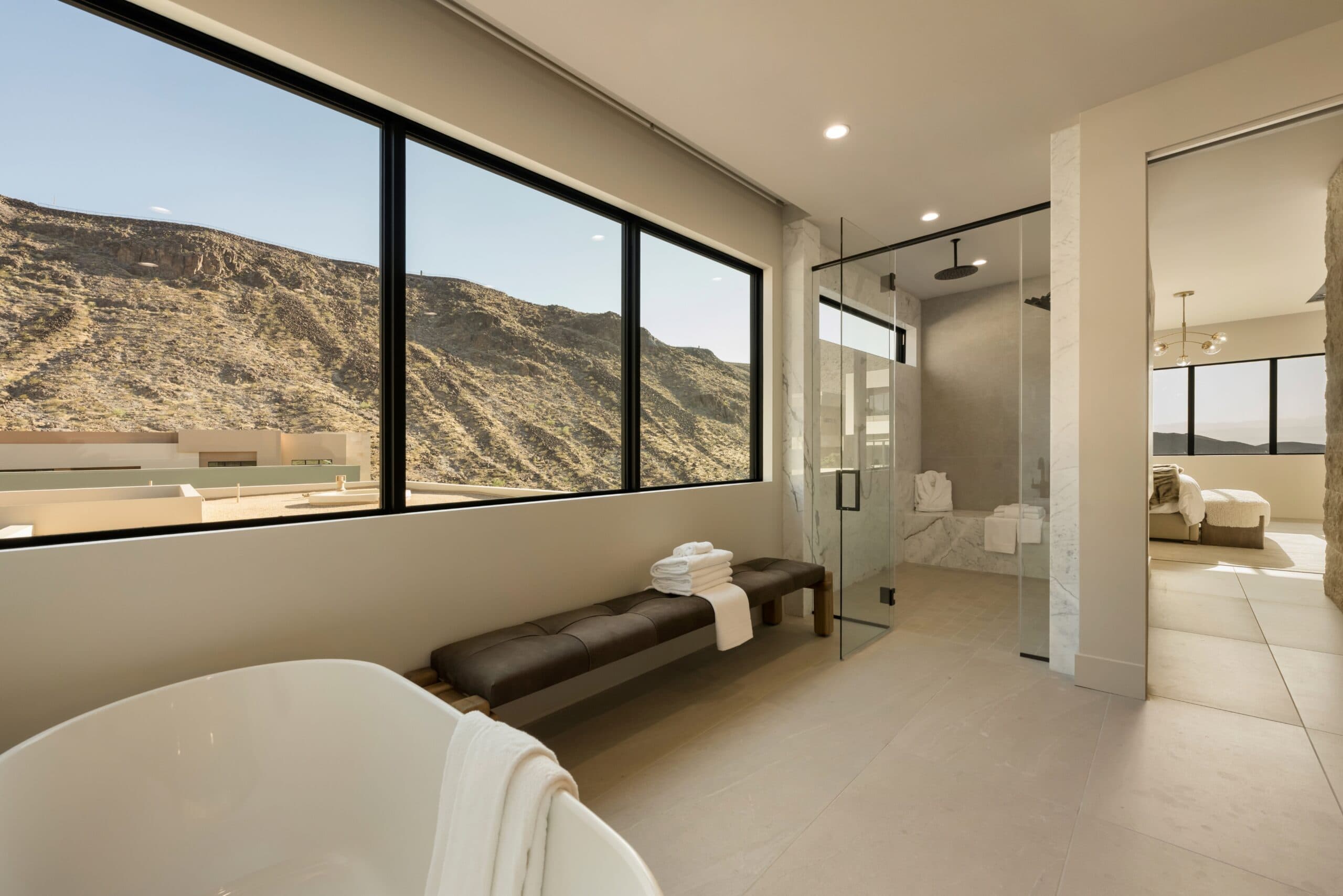 Luxury modern bathroom with a freestanding tub, glass-enclosed shower, expansive windows, and panoramic desert mountain views.