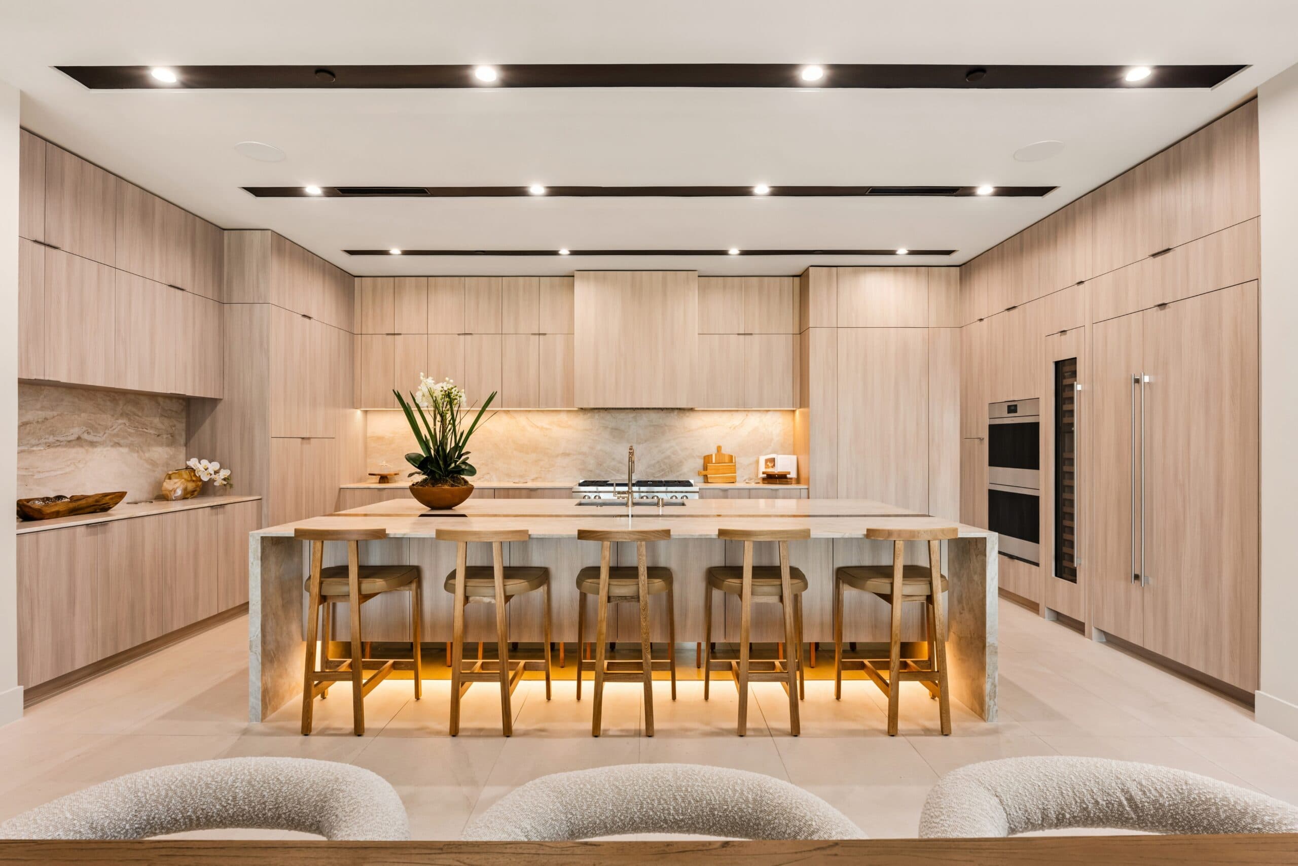 Luxury modern kitchen with light wood cabinetry, a large stone island, integrated appliances, and sleek contemporary lighting.