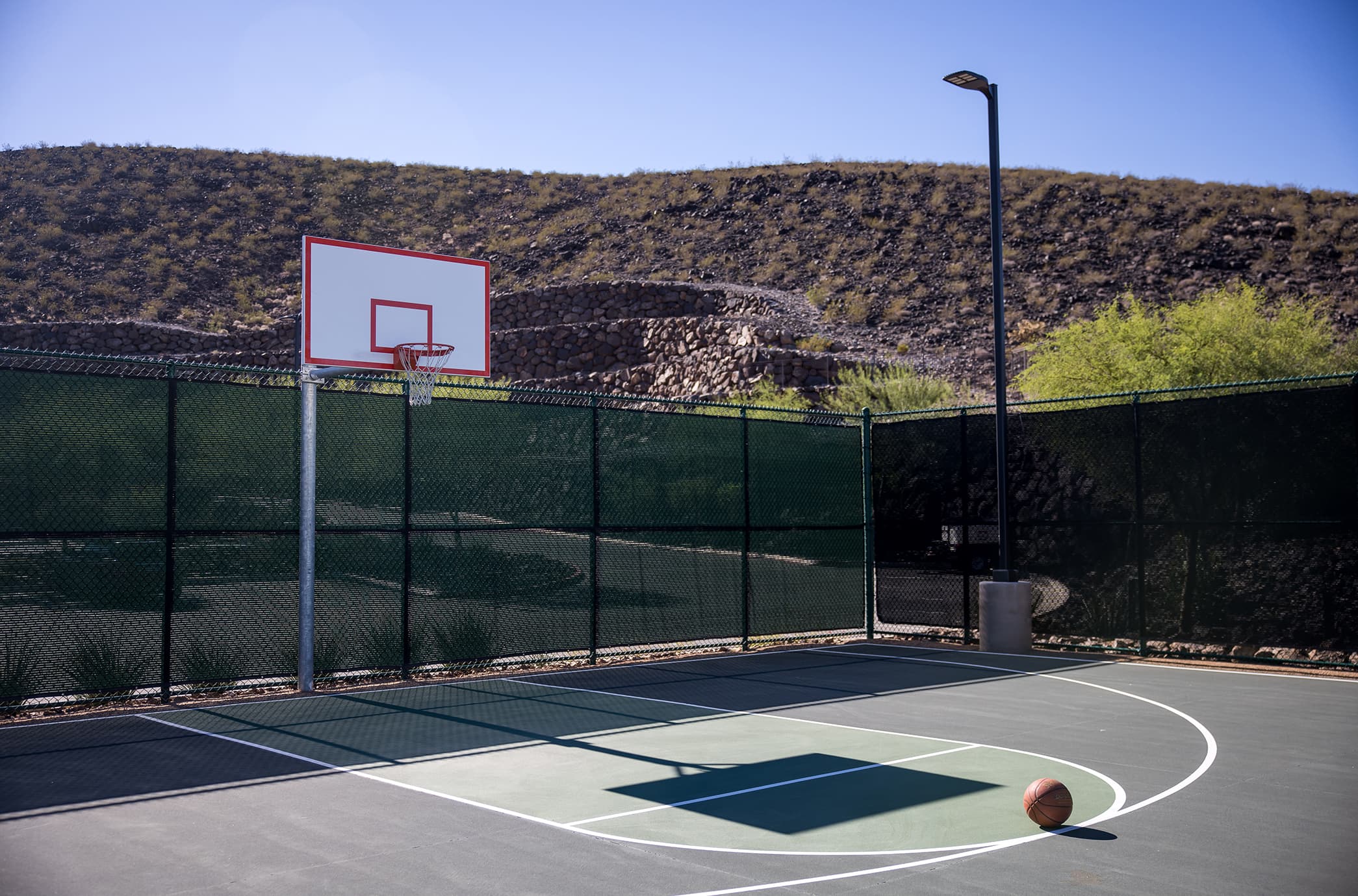 An outdoor half basketball court set against scenic desert and mountain views, featuring a single hoop, open play space, and surrounding fencing for recreation.