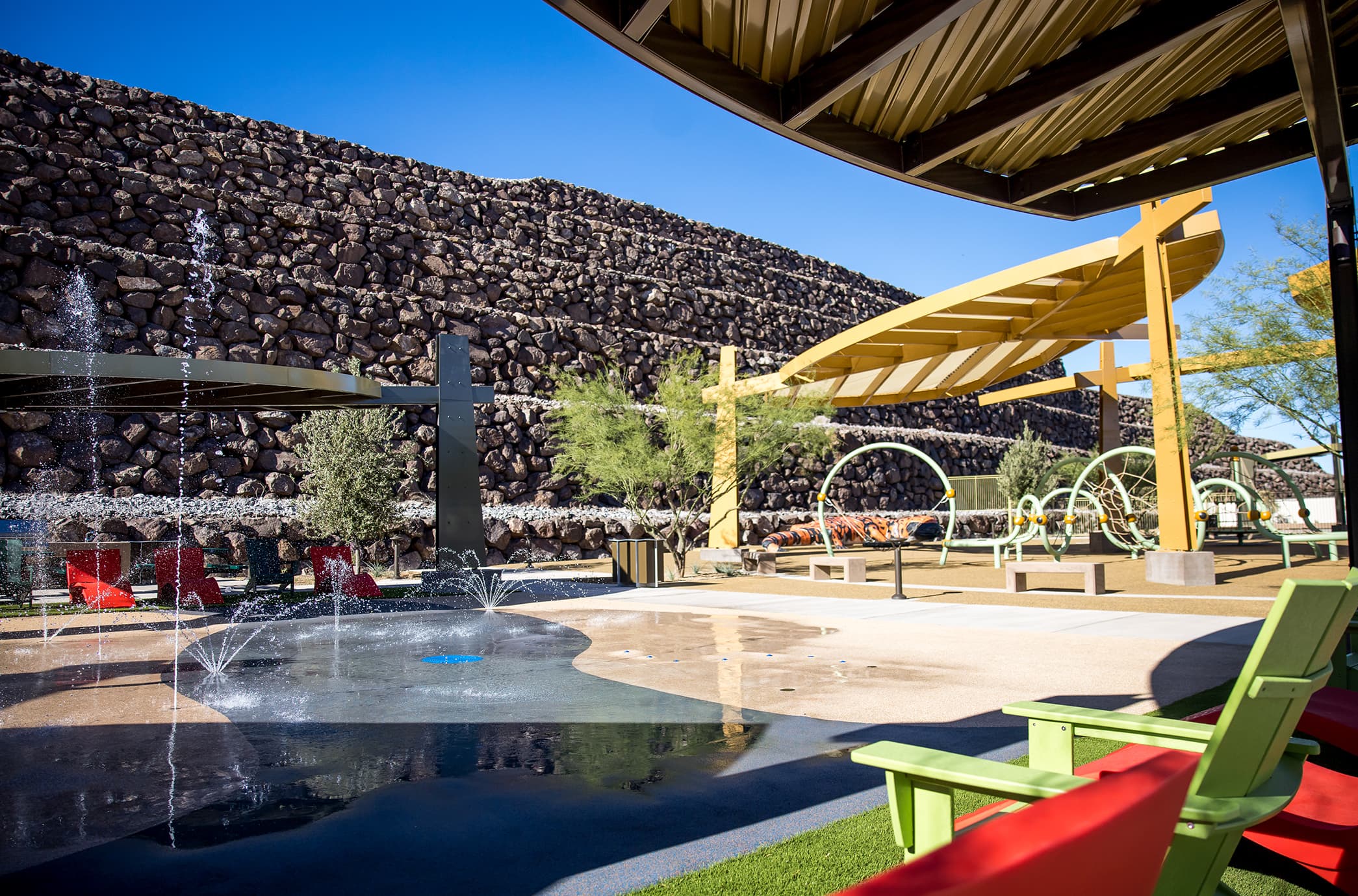 Family-friendly park at Ascaya featuring a shaded playground, splash pad with water features, and colorful seating set against terraced stone walls and desert landscaping.