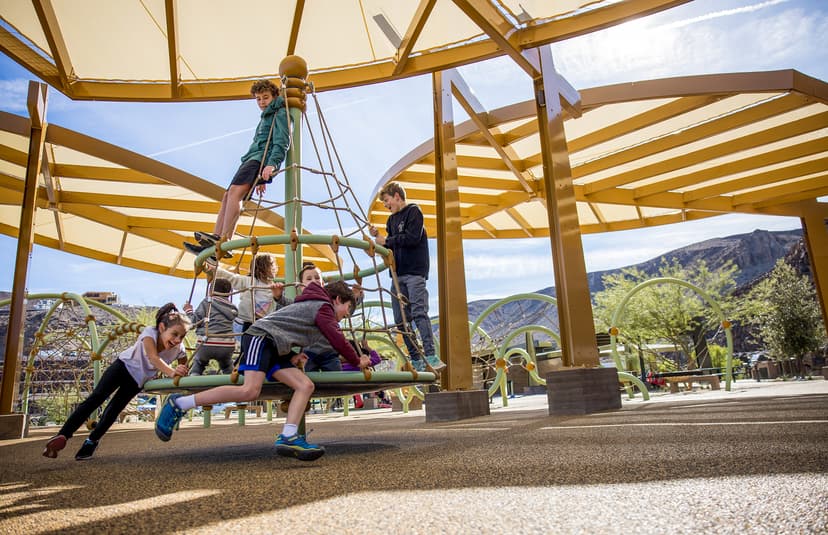Children playing on a modern playground with climbing structure, shaded canopy, and scenic mountain backdrop.