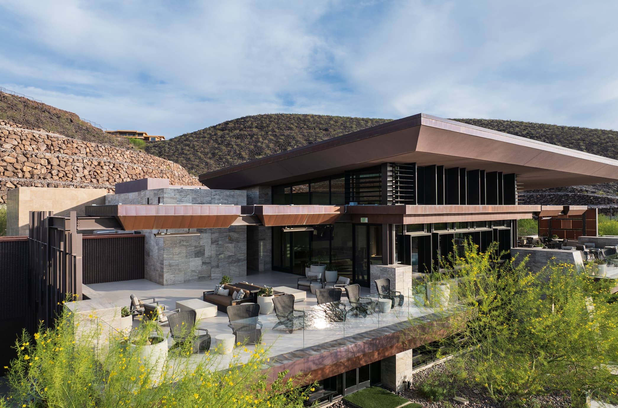 Modern luxury clubhouse exterior with rooftop terrace, contemporary architecture, and desert hillside surroundings.