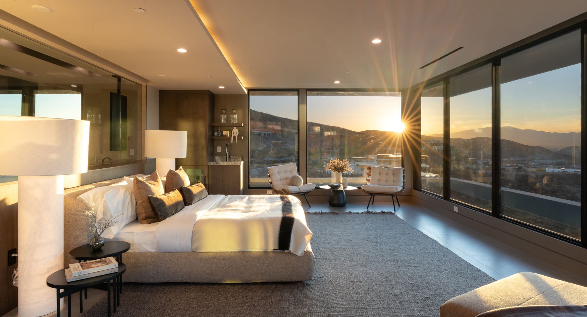 Serene contemporary bedroom retreat with floor-to-ceiling glass walls, warm minimalist finishes, and breathtaking sunset views across the surrounding desert mountains and Las Vegas valley.