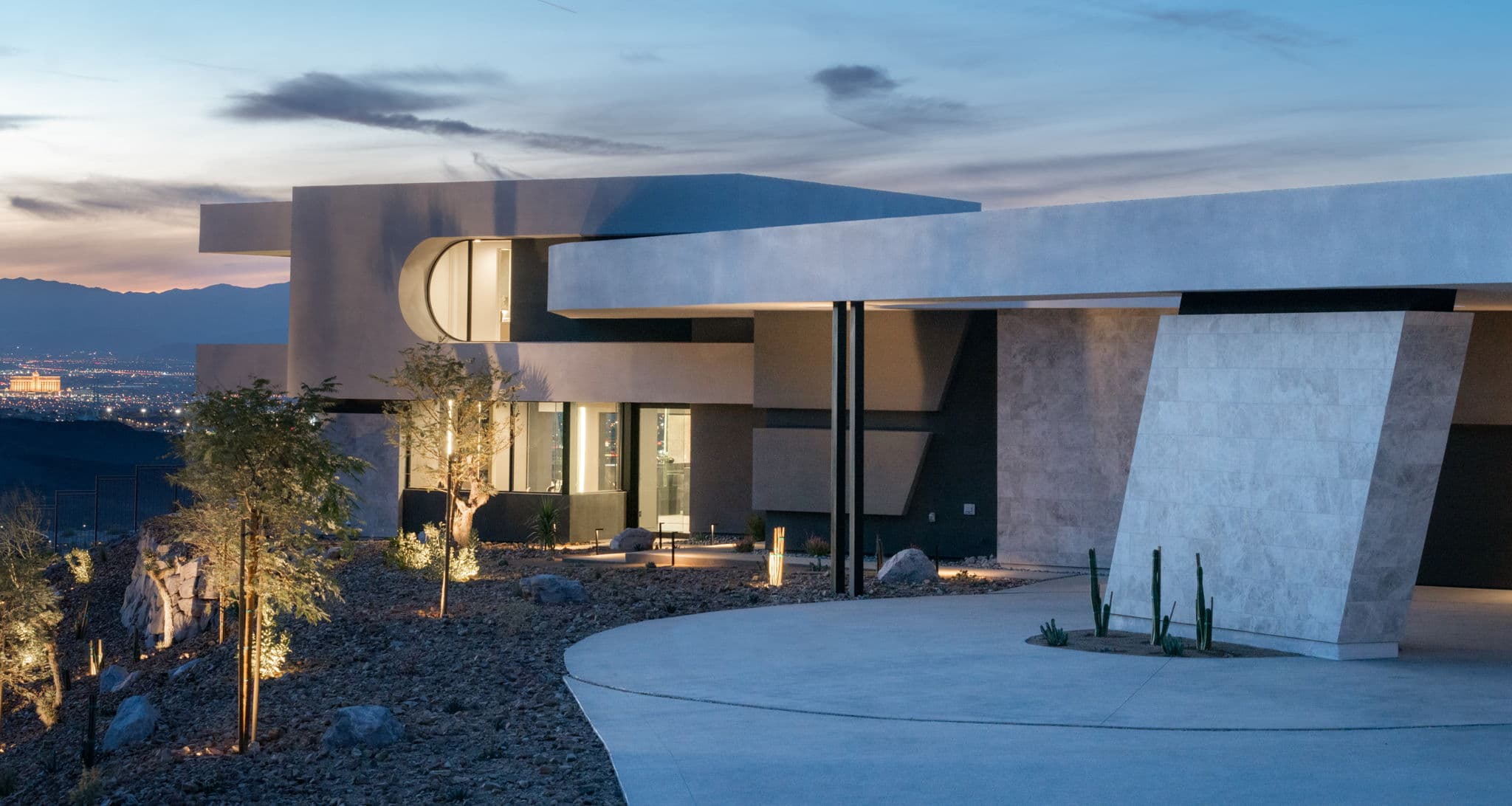 Contemporary luxury home exterior at dusk featuring bold modern architecture, dramatic geometric lines, desert landscaping, and panoramic mountain and city views illuminated by warm ambient lighting.