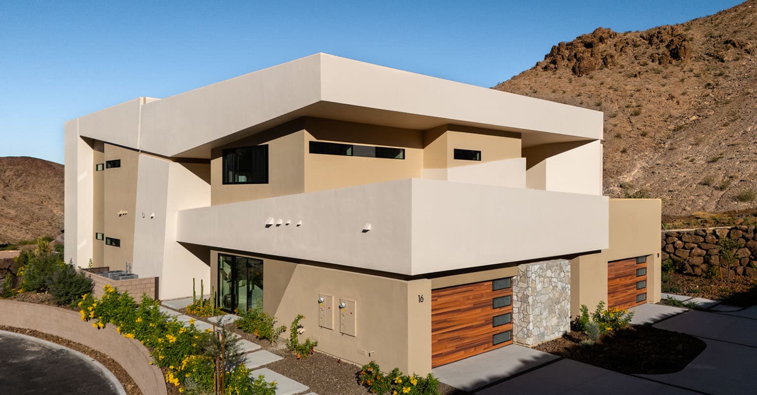 Modern desert luxury home exterior with clean architectural lines, flat roof design, wood garage doors, and scenic mountain backdrop.