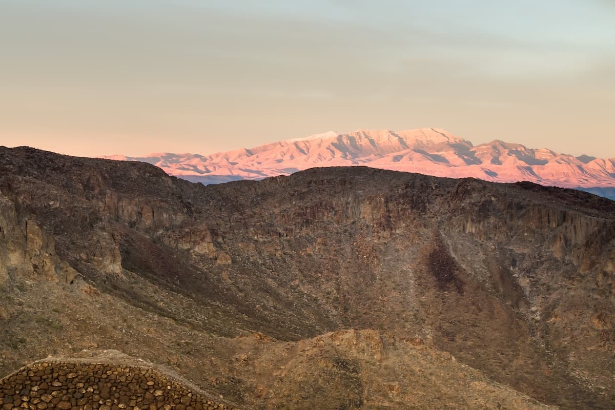 A rocky desert landscape at sunset with rugged hills in the foreground and a distant mountain range glowing pink under soft evening light.