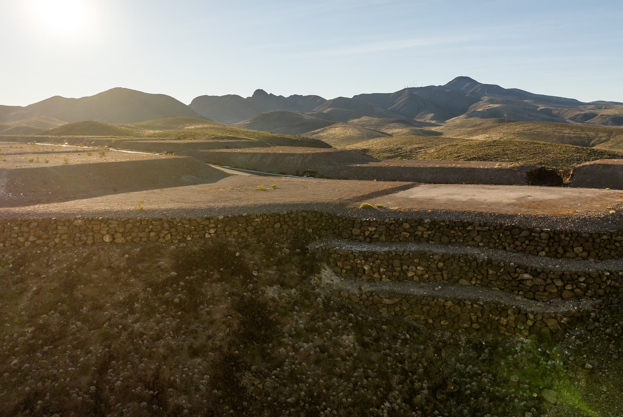 Aerial view of a desert hillside lot with a circular driveway pad overlooking a sprawling suburban city at sunset, with mountains in the distance.