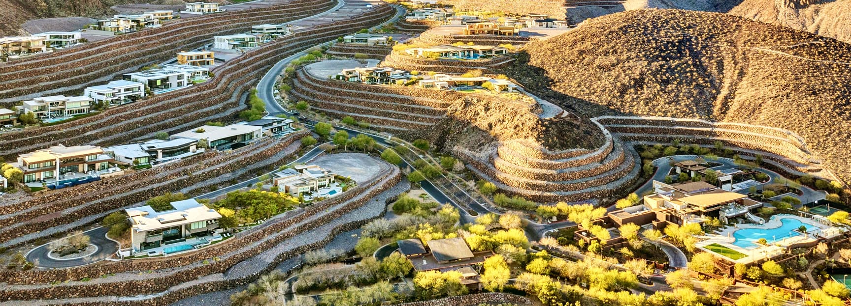 Aerial view of luxury desert hillside community with terraced homes, winding roads, and scenic mountain landscape.