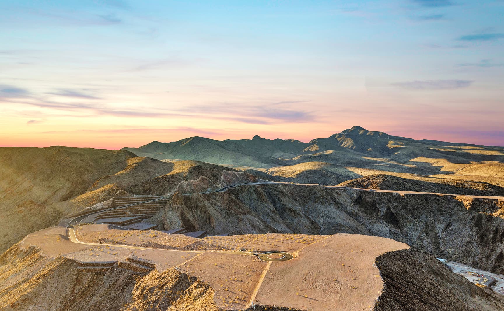 Aerial view of a desert hillside lot with a circular driveway pad overlooking a sprawling suburban city at sunset, with mountains in the distance.