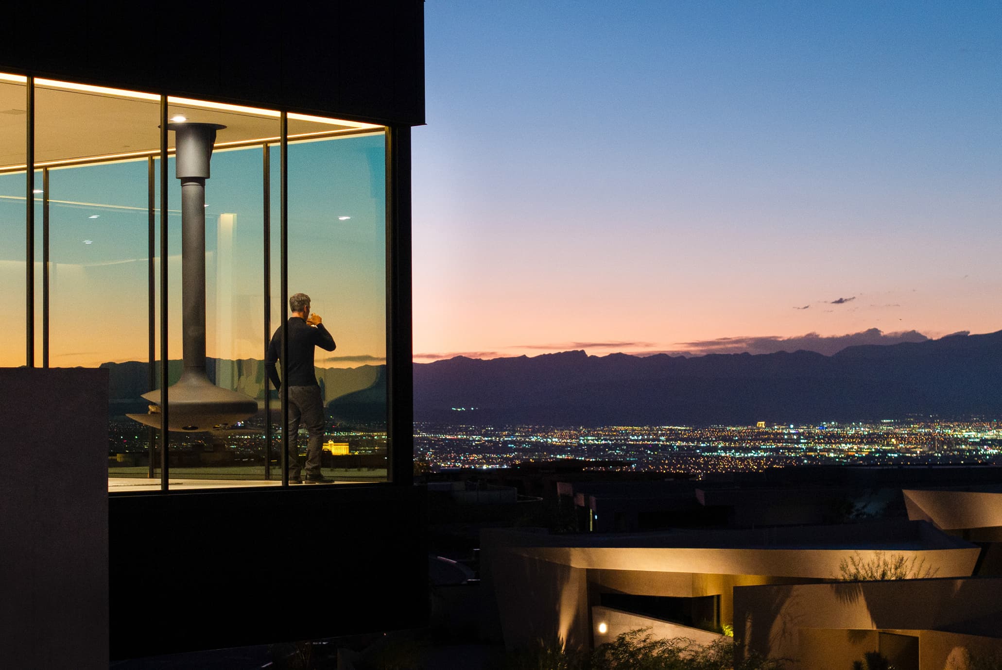 Man enjoying a drink inside modern luxury glass home overlooking city skyline at sunset.