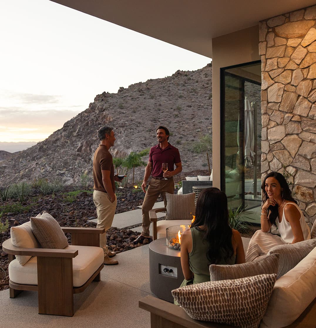 Friends enjoying drinks on a luxury patio with fire pit, modern seating, and desert mountain views at sunset.