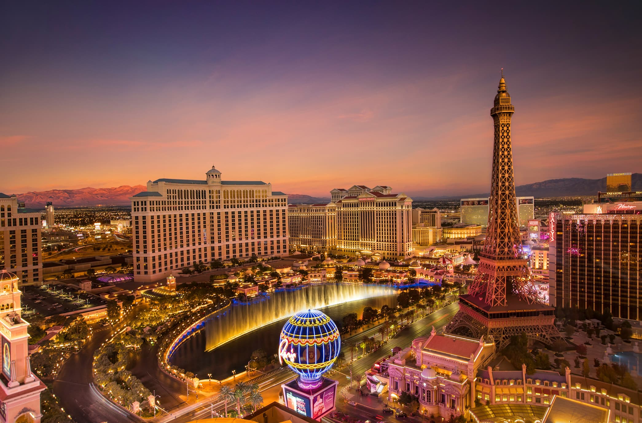 An iconic view of the Las Vegas Strip at sunset, featuring the Bellagio fountains, Paris Las Vegas Eiffel Tower, and vibrant city lights set against a desert mountain backdrop.
