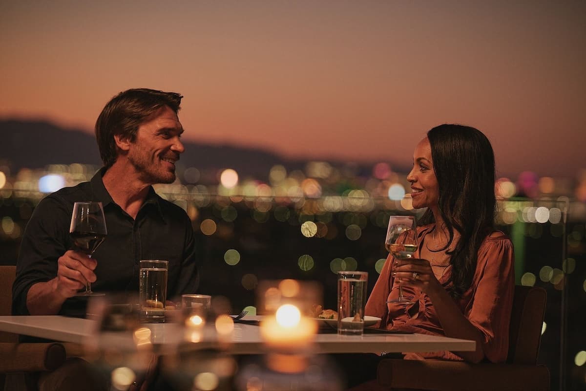 A man and woman sit at an outdoor table at sunset, smiling at each other while holding glasses of wine, with a city skyline glowing in the background.