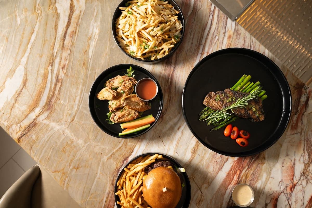 Grilled steak with asparagus, chicken wings with dipping sauce, burger with fries, and loaded fries on a marble table.