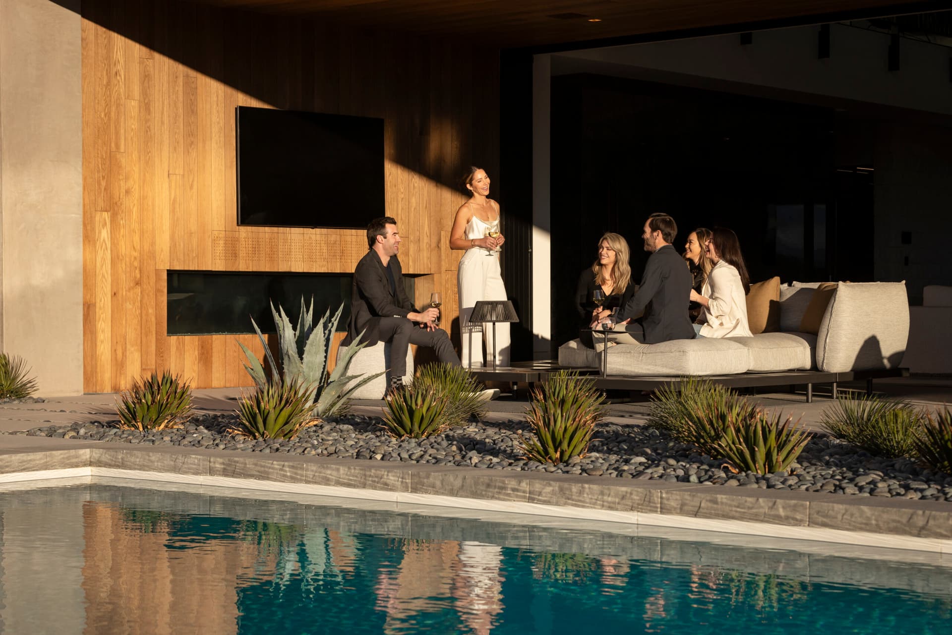 Group of friends relaxing by a modern poolside patio at a luxury home, enjoying drinks and conversation beside landscaped desert plants and a contemporary outdoor lounge.