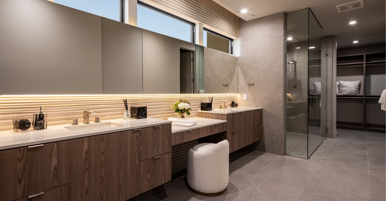 Contemporary spa-style bathroom featuring a long floating double vanity with wood cabinetry, integrated sinks, and a softly lit mirror, alongside a glass-enclosed shower and an open walk-in closet in a neutral, modern palette.