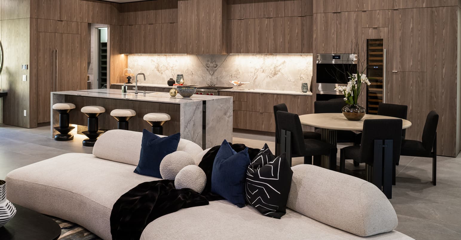 Open-concept living and dining area with a sculptural curved sofa, warm wood cabinetry, and a marble waterfall kitchen island with bar seating, complemented by soft ambient lighting and modern finishes.