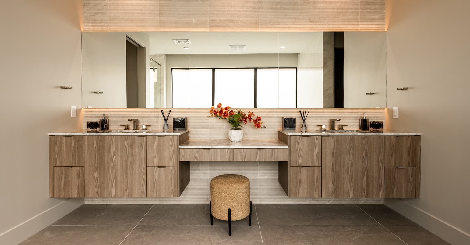 Modern double vanity bathroom with floating wood cabinetry, dual sinks, a large backlit mirror, and a central makeup station with a stool, accented by warm lighting and neutral stone finishes.