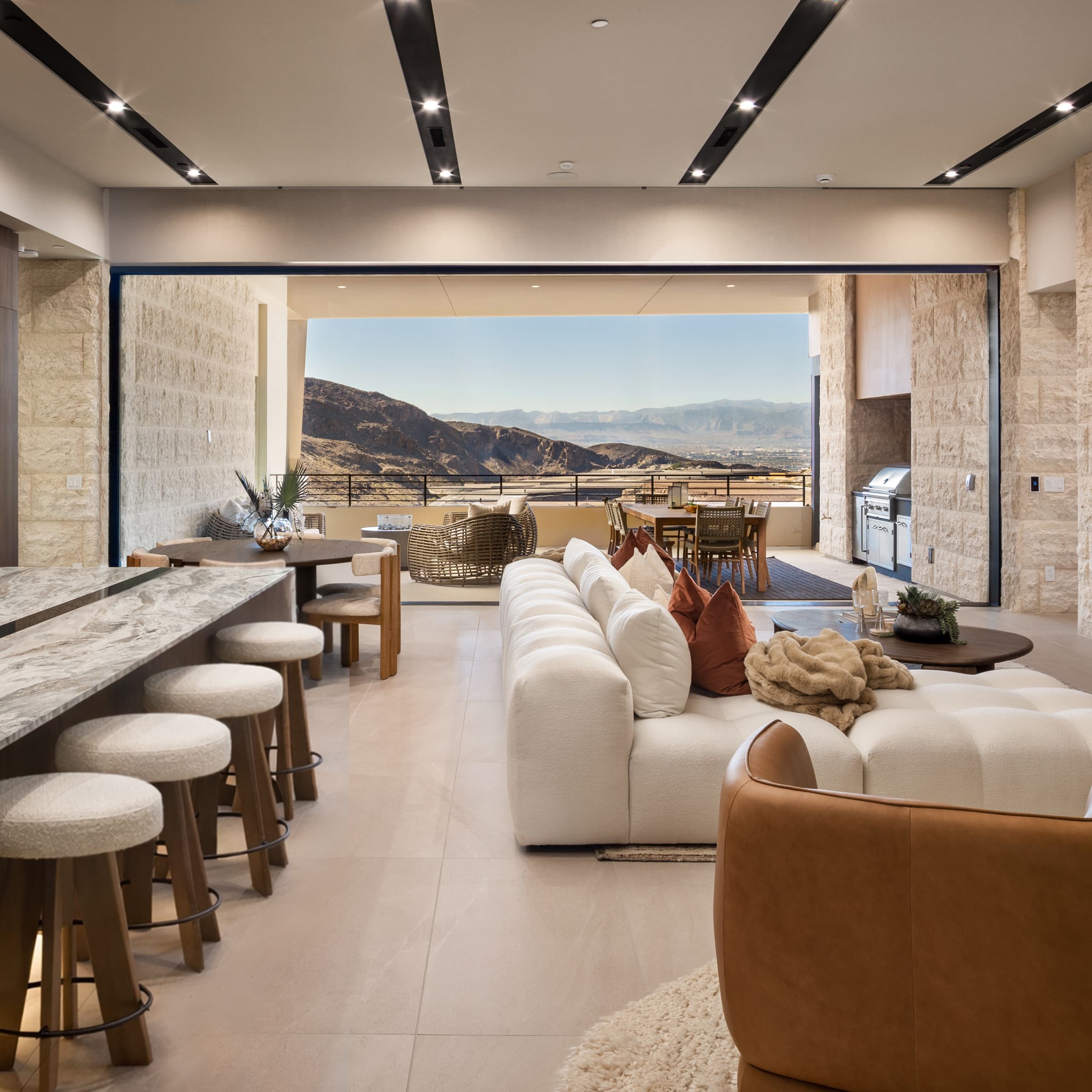 Open-concept living space with a marble island kitchen, bar seating, and plush lounge area, flowing seamlessly to a covered terrace with outdoor dining, built-in grill, and panoramic desert mountain views.