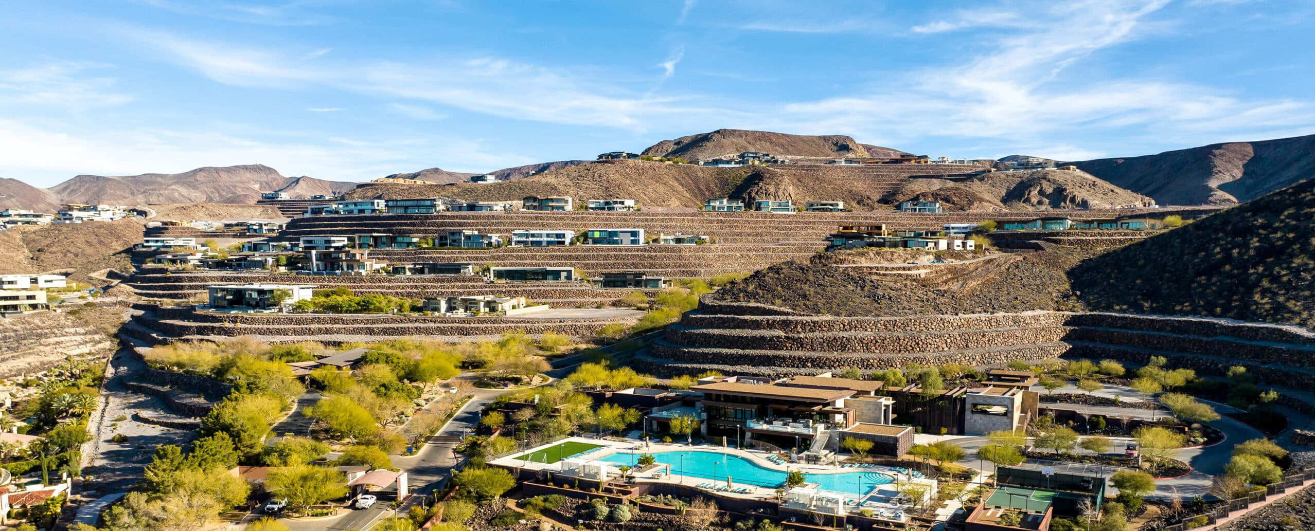 Aerial view of Ascaya’s terraced hillside community, showcasing modern luxury homes, the clubhouse, resort-style pool, tennis courts, and winding roads set against a dramatic desert landscape.