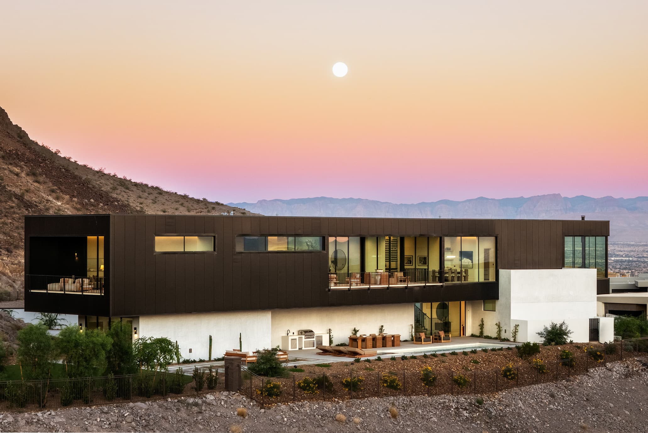 Modern custom home at Ascaya featuring sleek desert contemporary architecture, expansive glass walls, and seamless indoor-outdoor living set against a scenic mountain backdrop at sunset.