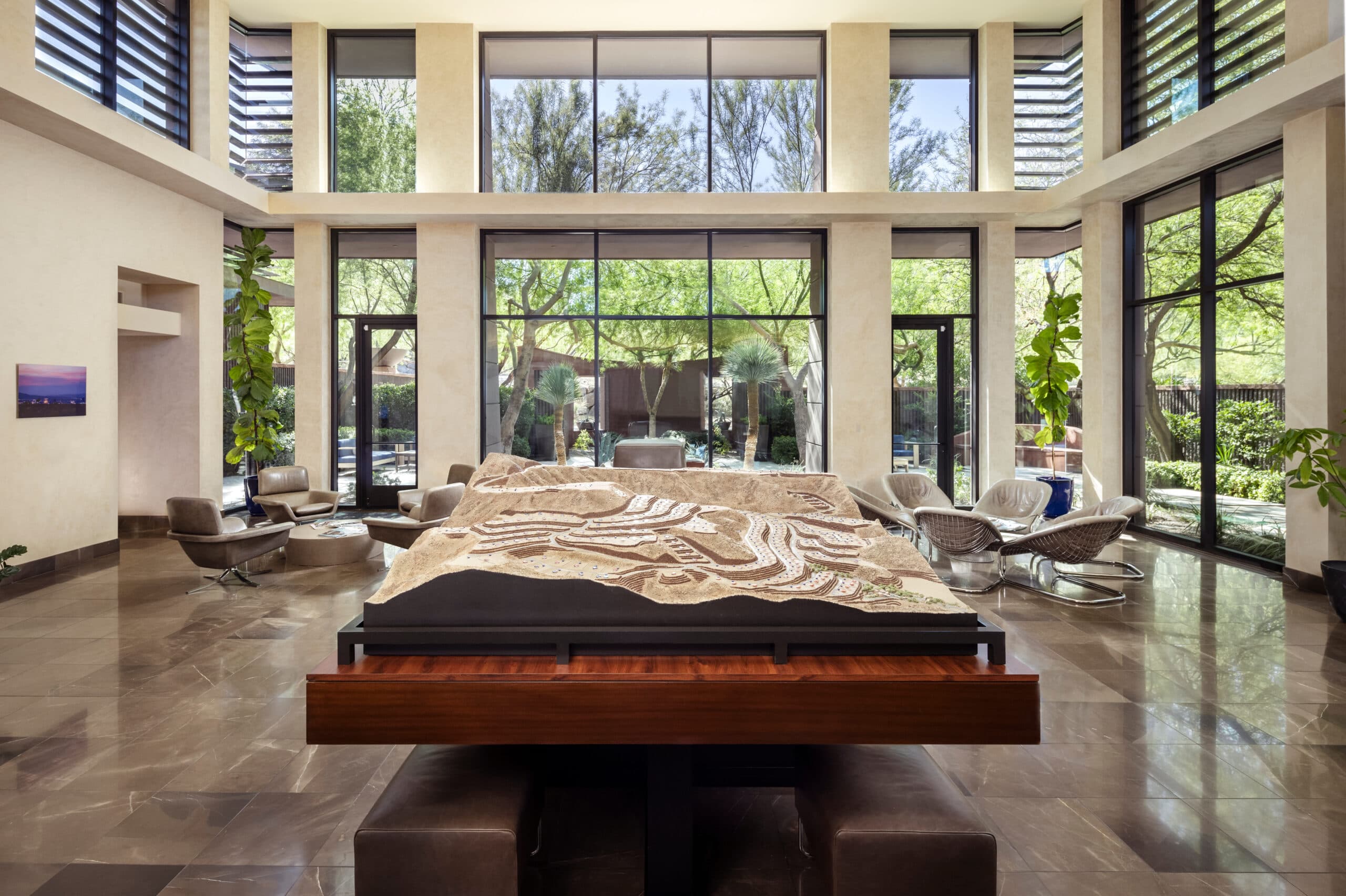 Modern luxury sales gallery interior with large architectural site model display, floor-to-ceiling windows, and landscaped courtyard views in a bright, open-concept space.